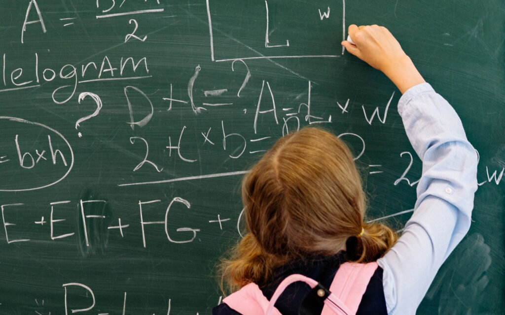Student with a pink backpack writes math equations on a green chalkboard, seen from behind in a classroom setting.