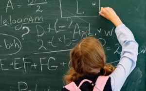 Student with a pink backpack writes math equations on a green chalkboard, seen from behind in a classroom setting.