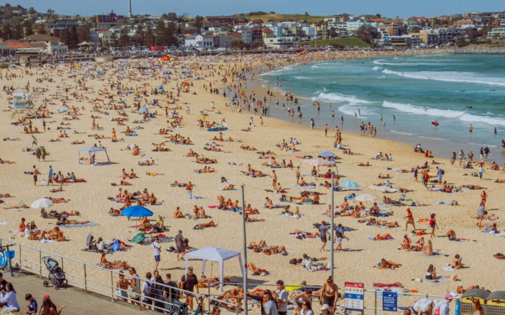 Crowded beach with sunbathers, umbrellas, and swimmers along a sandy shore, with buildings in the background.