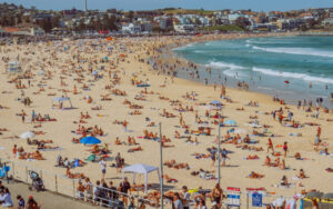 Crowded beach with sunbathers, umbrellas, and swimmers along a sandy shore, with buildings in the background.
