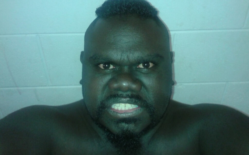 Close-up of a shirtless man with a mohawk and goatee making a fierce, grimacing expression at the camera against a pale wall.