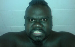 Close-up of a shirtless man with a mohawk and goatee making a fierce, grimacing expression at the camera against a pale wall.