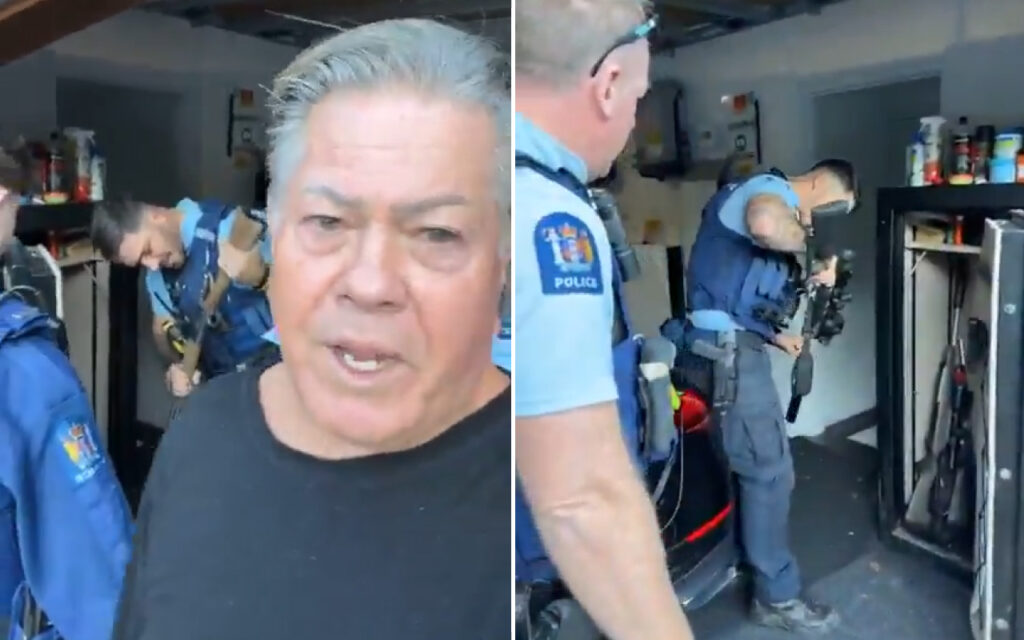 Close-up of a man in a black shirt speaking to the camera, with police officers and gear in a garage behind him.