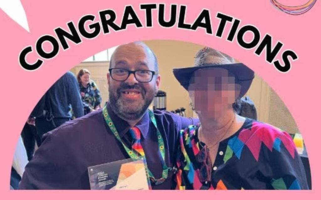 Two people pose for a celebration photo under a pink arch reading 'CONGRATULATIONS'; the man wears a purple shirt and colorful tie, the woman wears a black hat and a bright geometric top with her face blurred.