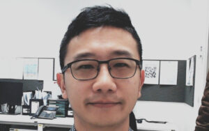 Close-up portrait of a man wearing black rectangular glasses in a bright office cubicle behind him is a desk with monitors and files.