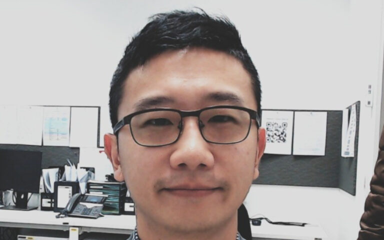 Close-up portrait of a man wearing black rectangular glasses in a bright office cubicle behind him is a desk with monitors and files.