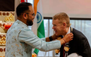 Two men share a joyful moment as one places a garland around the other's neck at a formal ceremony, with an Indian flag in the background.