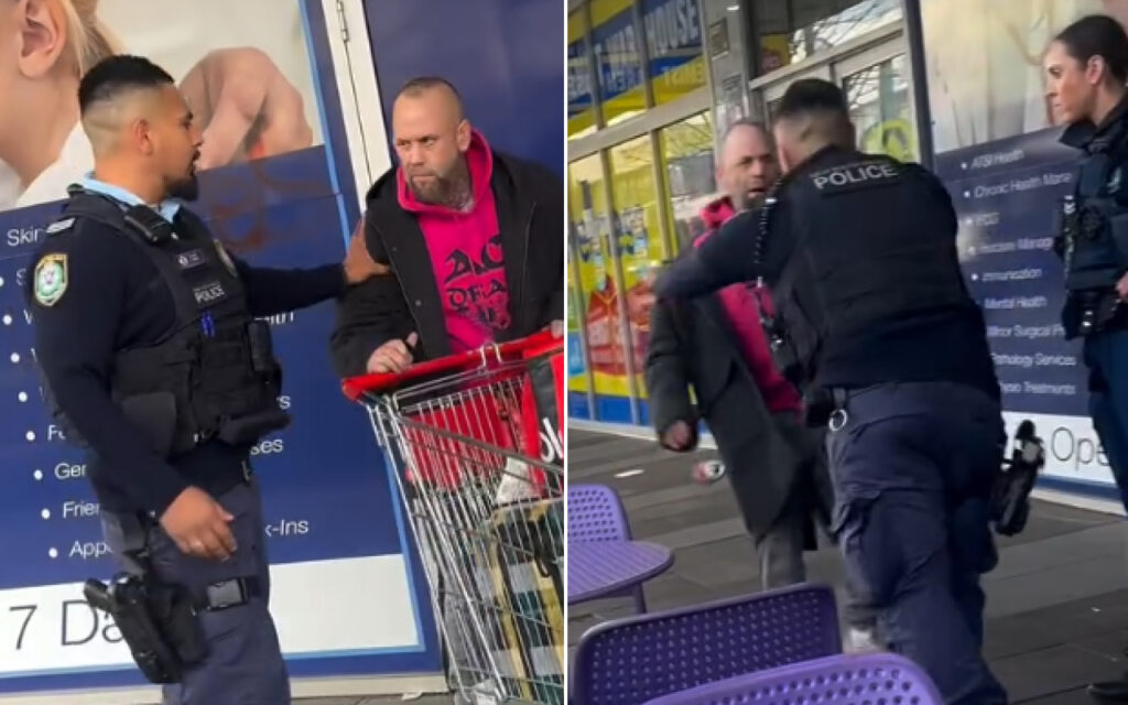 Two police officers confront a man with a red hoodie near a shopping cart outside a storefront.