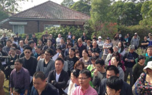 Large crowd of adults standing outdoors in a yard, attending a community event with a house in the background.
