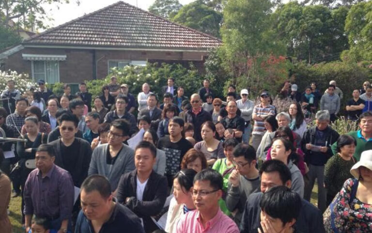 Large crowd of adults standing outdoors in a yard, attending a community event with a house in the background.