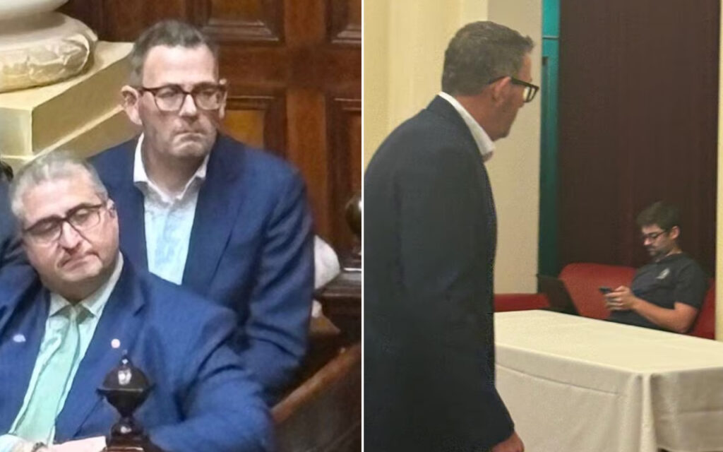Two men in blue suits sit in a formal room; one rests his head on his hand while the other stands behind him, both wearing glasses.