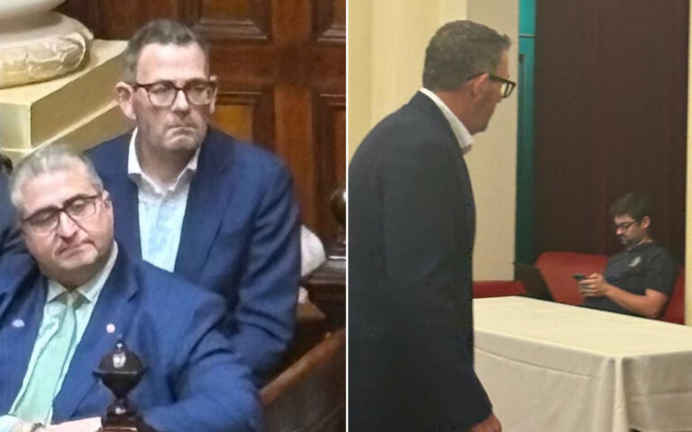 Two men in blue suits sit in a formal room; one rests his head on his hand while the other stands behind him, both wearing glasses.