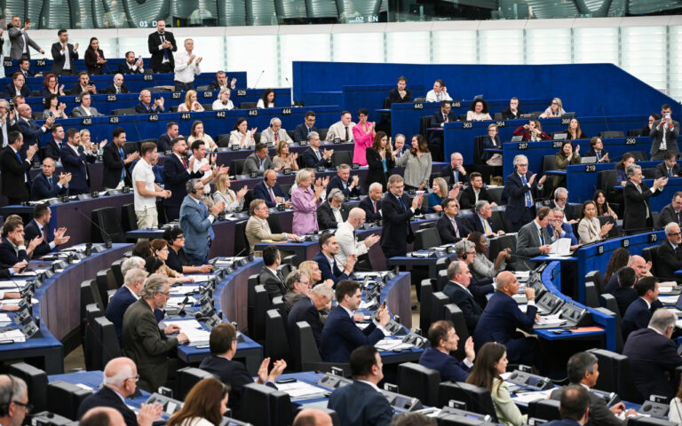 European Parliament chamber filled with politicians applauding during a session at blue desks and seating tiers