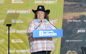 Older woman in a black cowboy hat speaks at a podium labeled Townsville Bulletin during a Bush Summit event, with yellow-green backdrop.