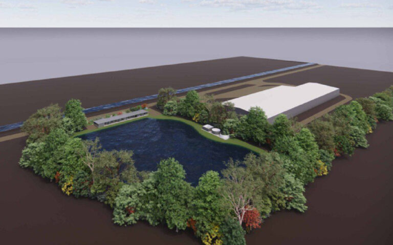 Aerial view of a wooded lakeside area with a dark blue pond, surrounding trees, a long rectangular building, and white storage tanks near the shore.