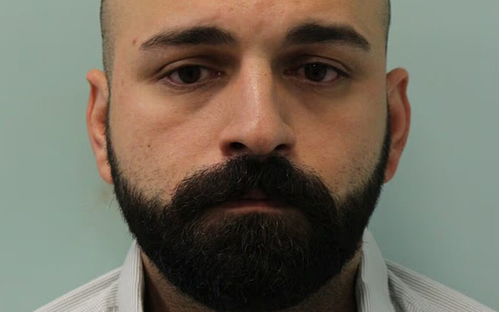 Close-up of a man with a thick black beard and mustache, wearing a light striped shirt, facing the camera with a neutral expression
