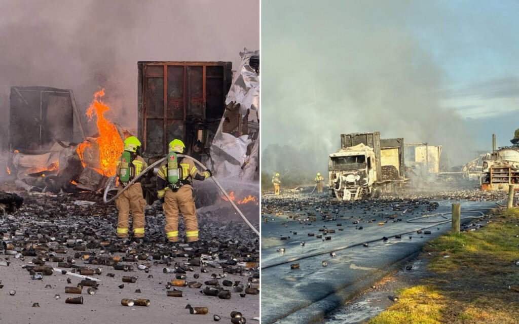 Two firefighters in yellow gear spray water at a burning wreck with scattered debris and containers.