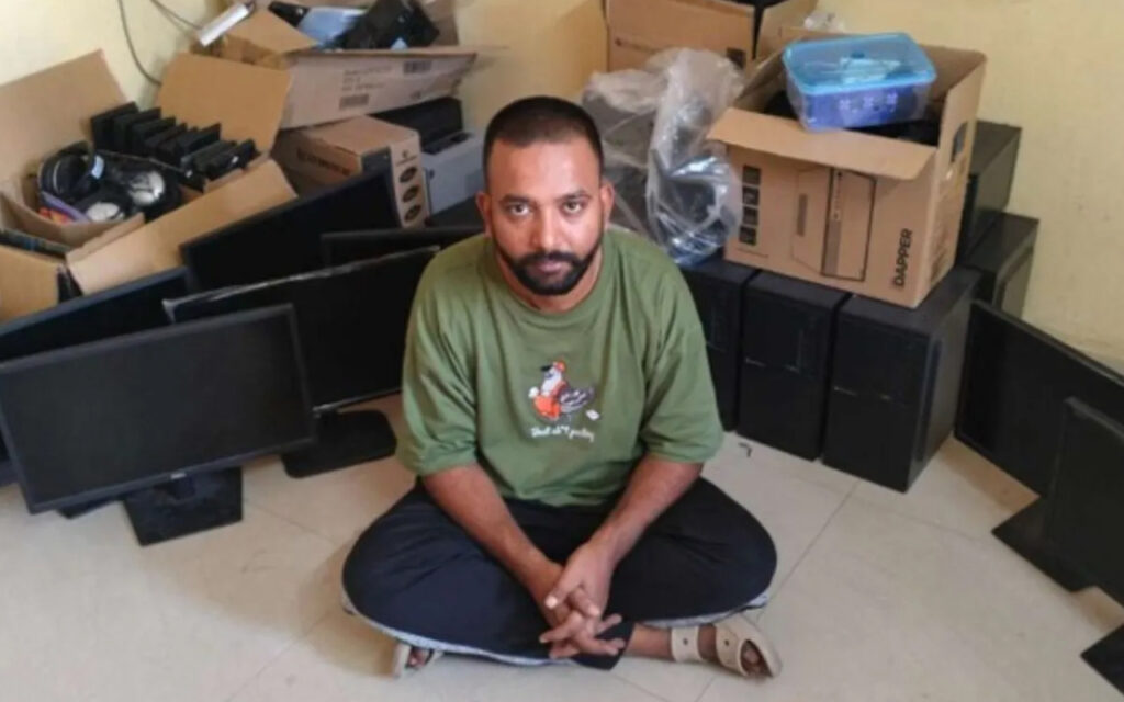 Man sits cross-legged on a tiled floor among stacked computer monitors and cardboard boxes in a cluttered room.