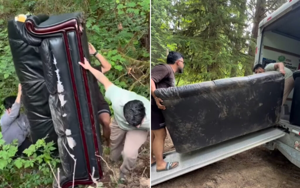 People carry a large black sofa through a wooded area toward a moving truck.