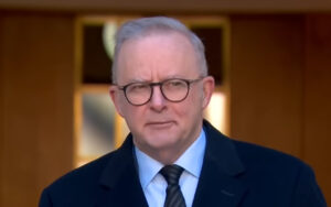 Man in a dark suit and blue shirt with a striped tie, wearing round glasses, standing in a warmly lit hallway.