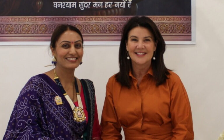 Two smiling women pose together indoors; one in a purple patterned sari with jewelry, the other in an orange blouse.
