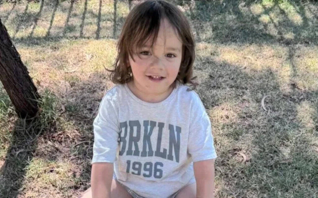 Smiling young child sitting outdoors on grass beside a tree, wearing a gray shirt with 'BRKLN 1996' in bold letters.