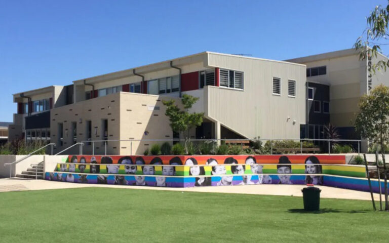 Modern school building with a rainbow-colored tiered mural of faces along the steps, green lawn in the foreground.