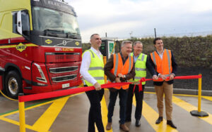 Five people in high-visibility vests cut a red ribbon beside a large red and yellow electric truck marked '100% ELECTRIC'.