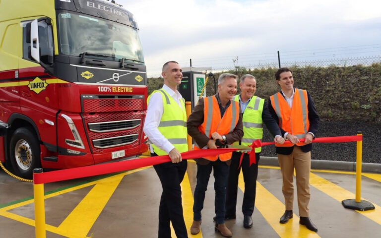 Five people in high-visibility vests cut a red ribbon beside a large red and yellow electric truck marked '100% ELECTRIC'.