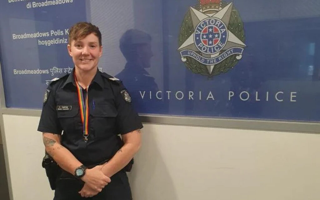 Female Victoria Police officer in uniform posing in front of a blue Victoria Police backdrop with a rainbow lanyard.