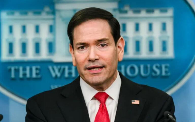 Man in a dark suit and red tie speaks at a White House briefing room backdrop, resembling a press briefing