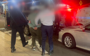 Detainee in handcuffs being escorted toward a police van at night, with officers and an ambulance nearby.