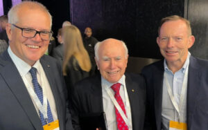 Three men in suits at a conference posing for a group photo, each wearing a lanyard and badge with a dark backdrop behind them.