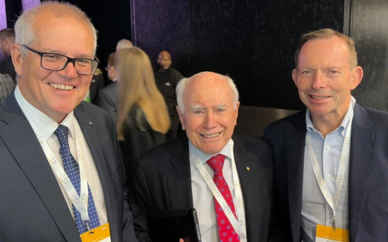 Three men in suits at a conference posing for a group photo, each wearing a lanyard and badge with a dark backdrop behind them.
