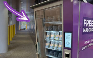 Purple naloxone vending machine in a hallway, with a touchscreen panel reading 'Touch screen to begin' on its side.