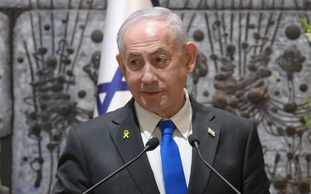 Older man in a dark suit and blue tie speaks into two microphones at a podium, wearing a yellow ribbon pin on his lapel with an Israeli flag in the background.