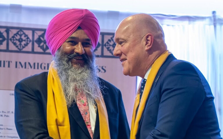 Two men in business attire greet each other at a formal event; one wears a pink turban and yellow scarf.
