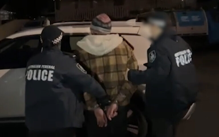 Two police officers arrest a man in a plaid jacket at night beside a car; hands cuffed behind his back.