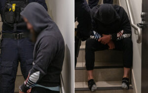 Hooded man with hands cuffed sits on stairs, an officer in tactical gear stands nearby in a doorway scene.