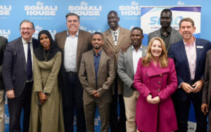 Group of ten diverse adults in business attire posing and smiling at a Somali House event, blue backdrop with logos.