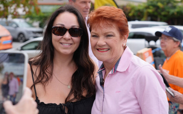 Two women posing for a selfie outdoors, one with dark hair and sunglasses and the other with short red hair in a pink polo, both smiling