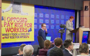 Panel discussion at a conference with a large yellow banner reading 'I opposed pay rise for workers...,' and a speaker at a podium.