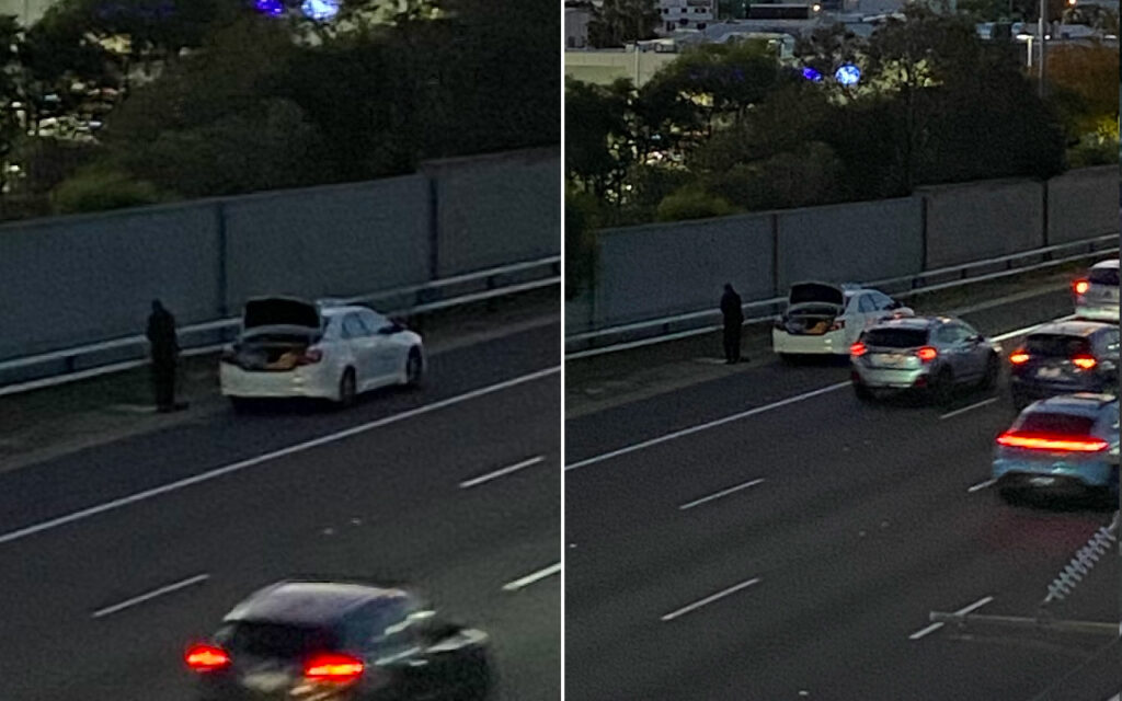 White sedan on the highway shoulder with the trunk open; a person stands nearby as traffic passes at dusk/sunset.