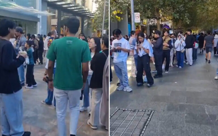 People queued along a city sidewalk on a sunny day, waiting outside storefronts; many are looking at phones or conversing.