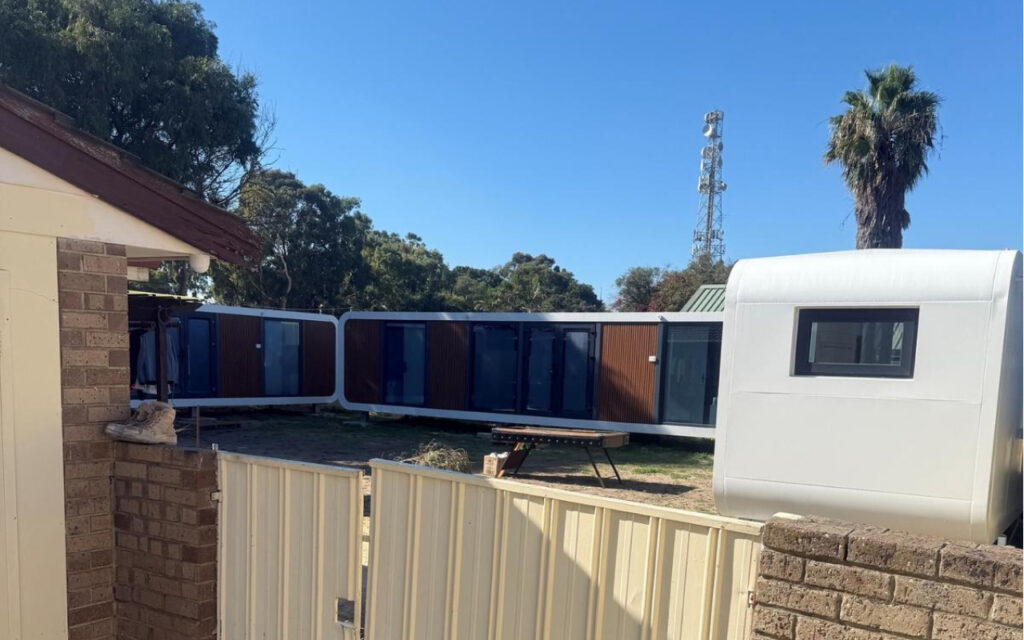 Outdoor view of connected blue modular units with a white curved trailer on the right and a palm tree against a clear blue sky.