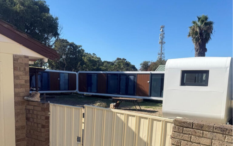 Outdoor view of connected blue modular units with a white curved trailer on the right and a palm tree against a clear blue sky.