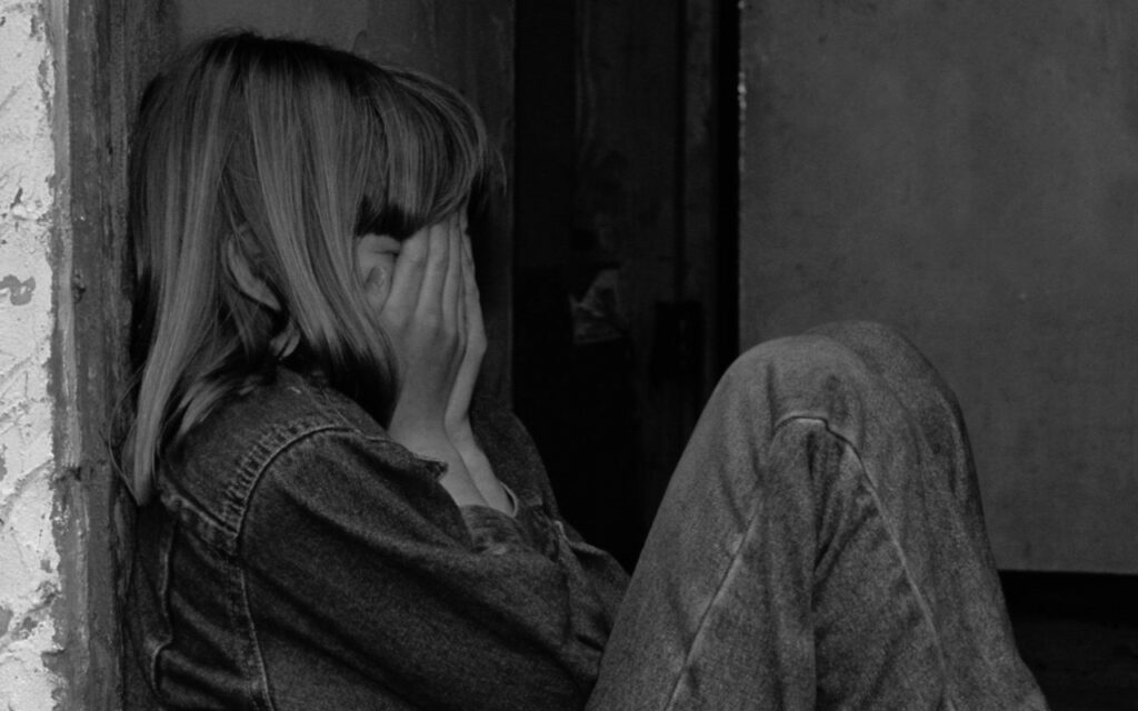 Person sitting against a wall with hands covering their face, clearly distressed or upset.