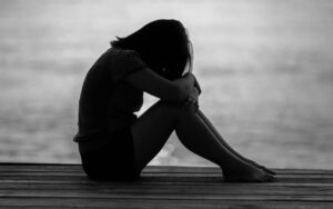 Silhouette of a person sitting on a wooden dock with knees drawn to chest, head bowed beside a body of water.