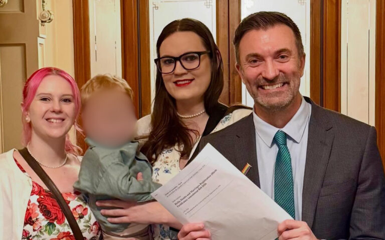 Four people pose for a photo indoors by wooden doors; a man in a gray suit and teal tie holds papers, flanked by a woman with pink hair and another woman wearing glasses, with a child whose face is blurred in the group photo.