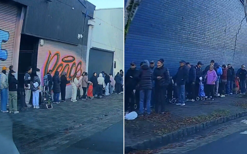 Crowd of people lined up along a city sidewalk beside a graffiti-covered wall in an urban street scene.